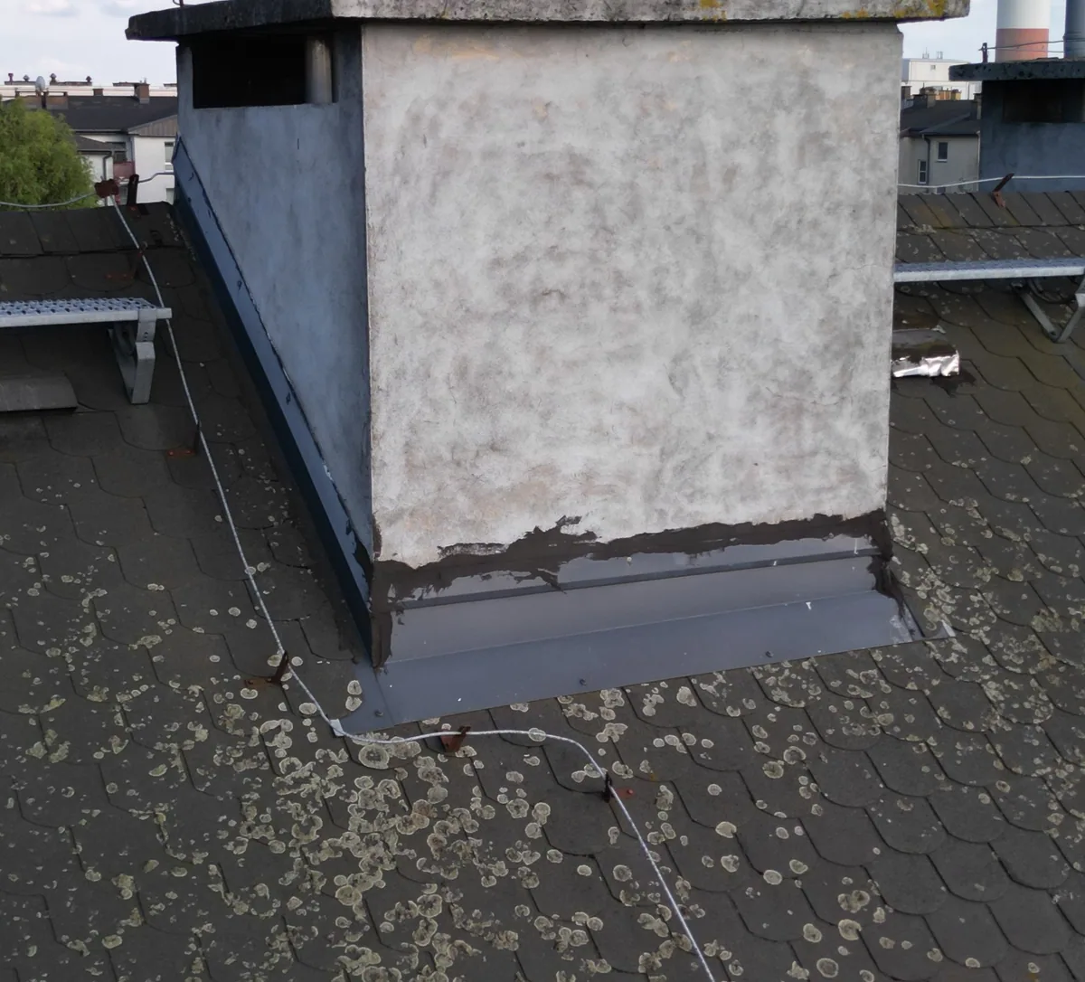Zbliżenie komina na skośnym dachu; u podstawy widoczne są metalowe obróbki i ciemny pas materiału uszczelniającego.
