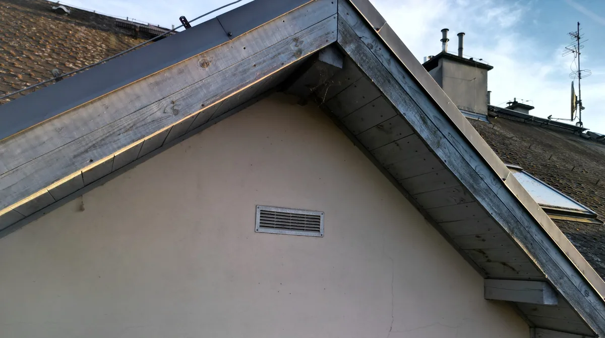 Szczyt budynku z szarymi deskami; na jasnym tynku pod szczytem widoczne są cienkie rysy.